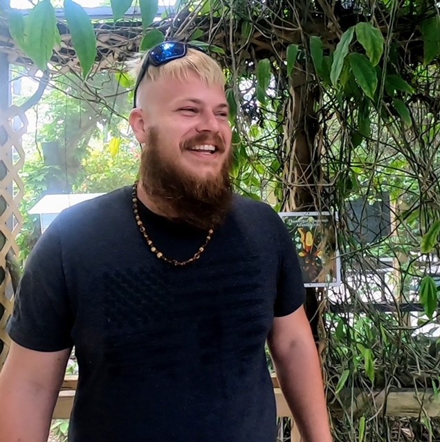 A man with blonde hair and beard standing in front of trees.