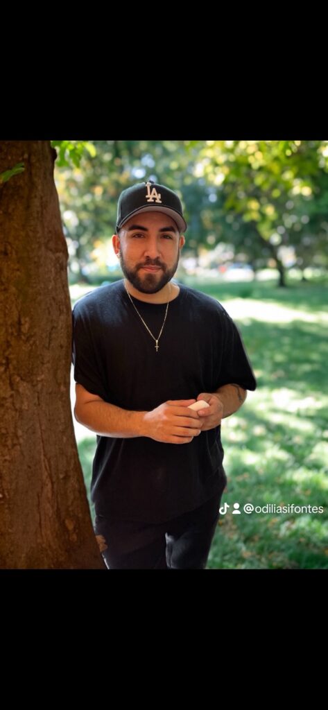 Man leaning on a tree in a park, wearing a black shirt and cap.
