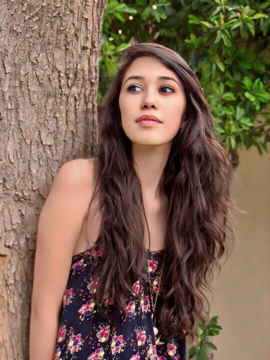 Young woman with long hair standing by a tree, looking thoughtful.