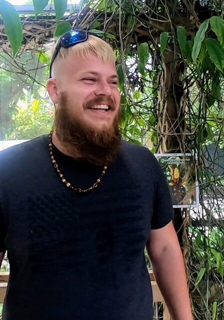 A man with blonde hair and beard standing in front of trees.