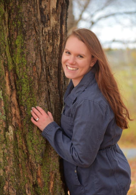 A woman leaning against a tree smiling for the camera.