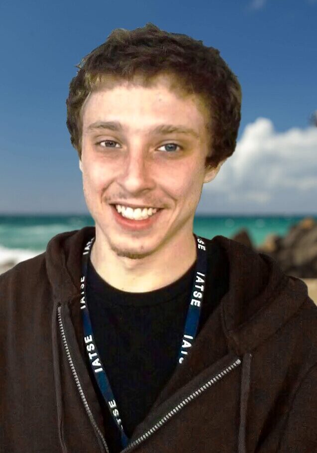 geoff-beachbackground A man with brown jacket and blue necklace standing on beach.